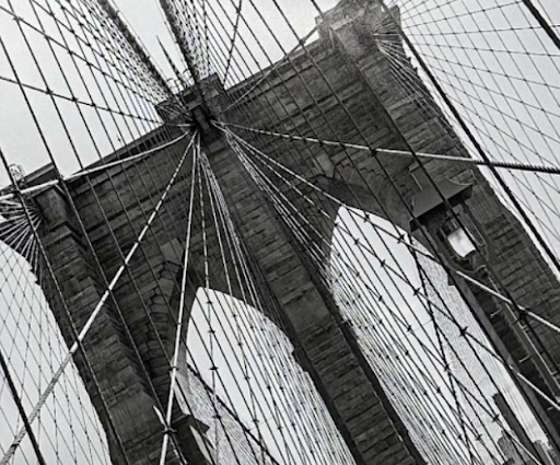 A detailed view of a bridge and suspension cables. The photograph is taken from a low angle, looking up at one of the bridge's towers and the intricate web of suspension cables that fan out from it. The stone tower is prominent, with its Gothic-style arches and the cables creating a geometric pattern against the sky.