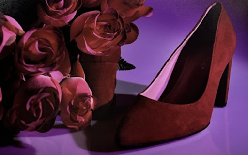 A pair of red high-heeled shoes placed next to a bouquet of red roses. The background is a gradient of purple shades, creating a striking contrast with the red color of the shoes and roses. The image stands out due to its artistic composition and the use of complementary colors to highlight the subjects.