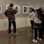 Four students observe a student photography showcase at Pellissippi State Community College. They stand in front of a beige wall adorned with black-and-white and color portraits, engaging with the displayed artwork. One student in a red plaid shirt gestures towards a photograph while the others listen attentively.