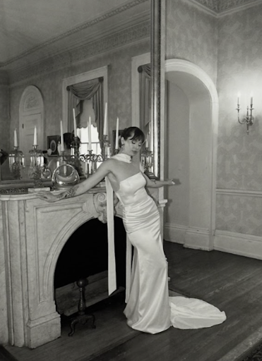 A black-and-white photograph of a woman in an elegant, form-fitting satin gown with a halter neckline and a long train. She leans gracefully against a marble fireplace in a historic, ornately decorated room, with tall candles and vintage décor adorning the mantel. Her eyes are closed, and her pose is relaxed yet poised, evoking a timeless, old-Hollywood glamour. The room features intricate wallpaper, classic architectural details, and a large mirror reflecting the sophisticated setting.