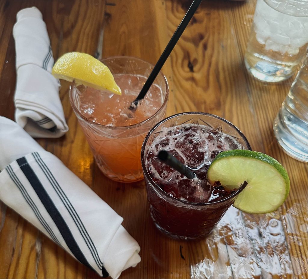 Two colorful non-alcoholic mocktails served on a wooden table at Tazza Kitchen in Richmond, Virginia. The drinks, "True Story" and "Sunday Best," are in short glasses with ice and garnishes—one with a lemon wedge and the other with a lime slice. Black straws are in both glasses, and white cloth napkins with black stripes are folded nearby. A glass of ice water is visible in the background.