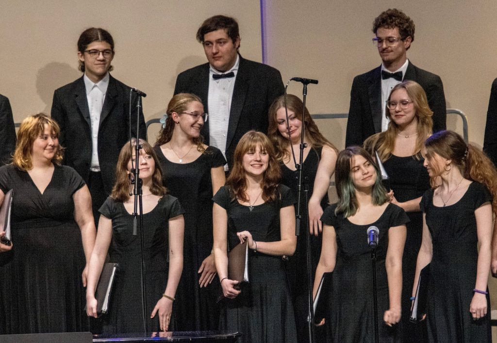 Photo of young women in black dresses preparing to sing in a choir concert. Three young men stand on risers behind them.