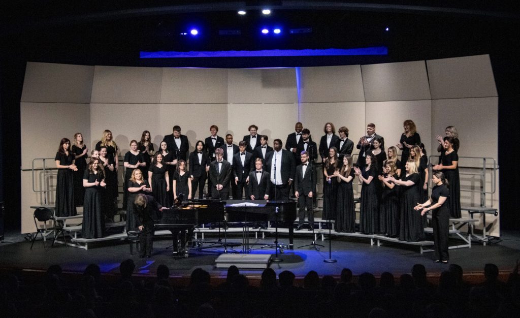 A full choir ensemble of young men and women stand on risers singing.