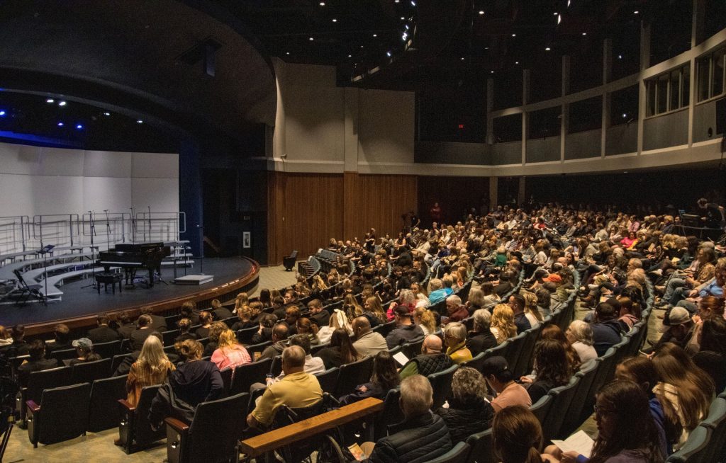 A photo of an audience looking at an empty stage with a grand piano on it.