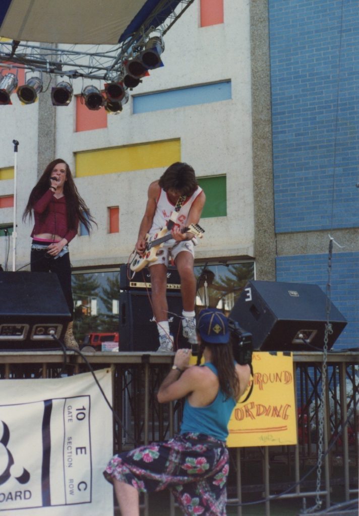 Photo of a band on a stage with a singer holding a microphone and guitarist playing.